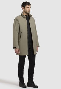 Beige vandtæt frakke med høj krave, frontknaplukning, sidelommer og sorte detaljer. Båret over en sort skjorte og sorte bukser.