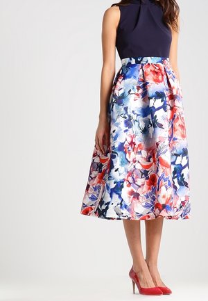 Ärmelloses marineblaues Top kombiniert mit einem plissierten Midirock mit hoher Taille mit rotem, blauem und weißem Blumenmuster und roten High Heels.