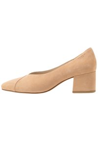 Beige mockasin med blockklack och spetsig tå, subtila sömnadsdetaljer och slät yta. Klackhöjd är måttlig och kraftig.