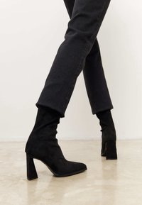 Black ankle boots made of suede with a high block heel and square toe, featuring a form-fitting design and smooth texture.