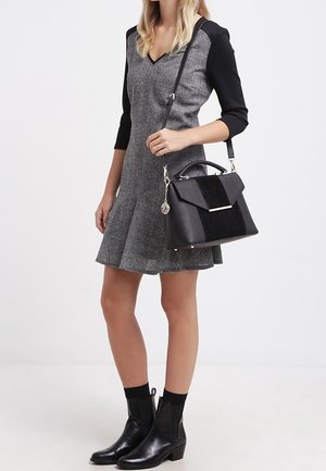 Mujer vestida con un vestido gris con mangas negras, botas negras hasta el tobillo y llevando un bolso negro con un adorno plateado en la correa del hombro.