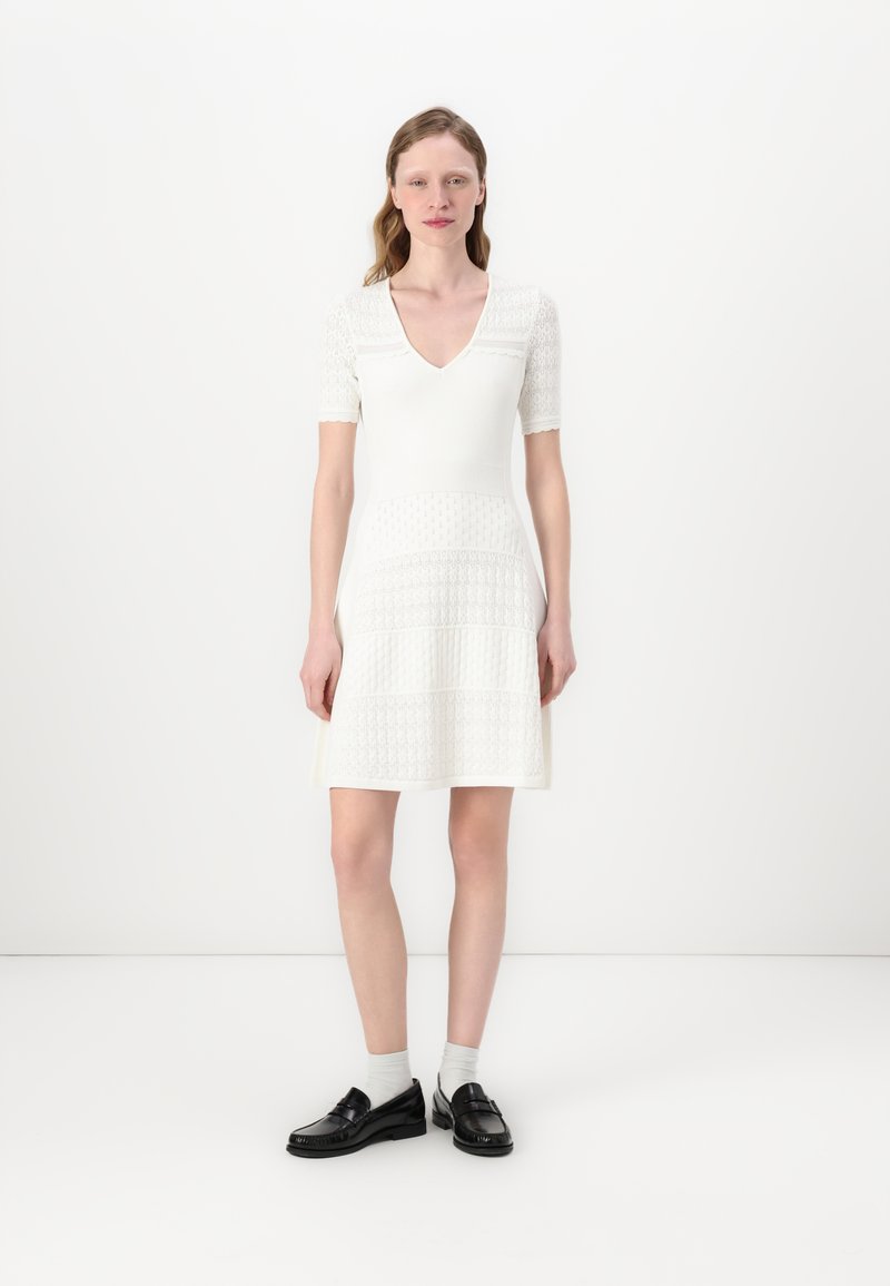 White short-sleeve dress with a V-neck, textured knit design, fitted bodice, and flared skirt. Paired with black loafers and gray socks.