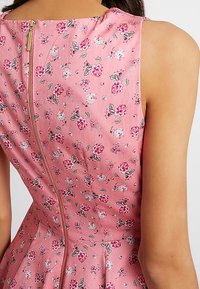 Zadní pohled na bezrukávové růžové šaty s malým květinovým vzorem a viditelným zlatým zipem, který probíhá středem.