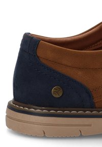 Zapato de ante marrón y azul marino con superficie texturizada, forma redondeada, acentos de cuero marrón y un logo circular de latón en el lateral.