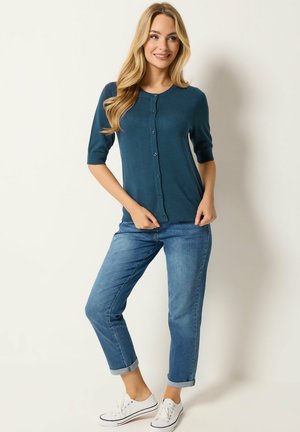 Kvinde med langt blondt hår iført en knappet teal cardigan, opkrogede blå jeans og hvide sneakers, stående mod en lys baggrund.