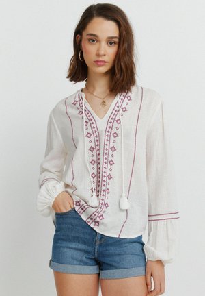 Kvinde iført hvid bluse med lilla broderi og kvaster, kombineret med opkrøjede denimshorts, stående med den ene hånd i lommen.