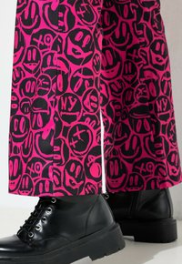 Roze broek met een zwart smiley-gezicht patroon met verschillende gezichtsuitdrukkingen. Gepaarde met zwarte veterschoenen met een stevige zool.