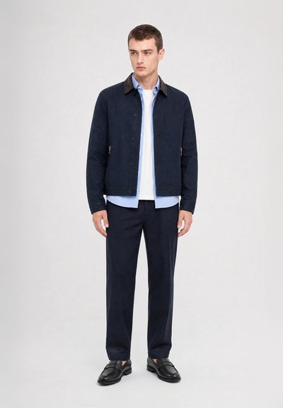 Jeune homme portant une veste bleu marine sur une chemise bleu clair et un t-shirt blanc, un pantalon bleu marine et des mocassins en cuir noir, debout devant un fond blanc.