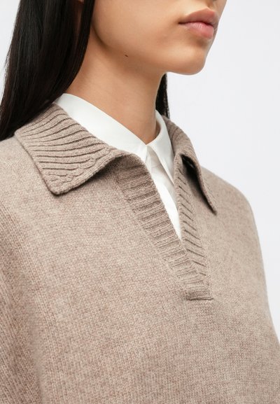 Beež kudumitega kampsun, millel on soonikkoes krae ja V-kaelus, kantud valge kraega särgi peal. Pehme tekstuur ja õmblusteta viimistlus.