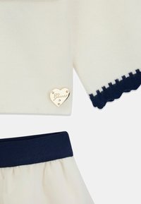 Haut en maille blanc avec ourlet festonné bleu marine, orné du logo Guess en forme de cœur doré. Jupe en tulle crème assortie avec ceinture élastique bleu marine.