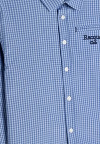 Camisa a cuadros azul y blanca con cuello abotonado, que cuenta con un bolsillo en el pecho y un logo bordado de "Racquet Club". Botones blancos.
