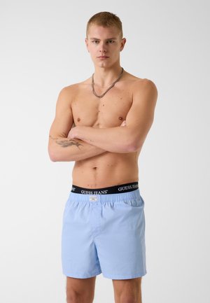Jeune homme torse nu aux cheveux courts, les bras croisés, portant un short bleu clair et une ceinture noire avec le texte "GUESS JEANS".