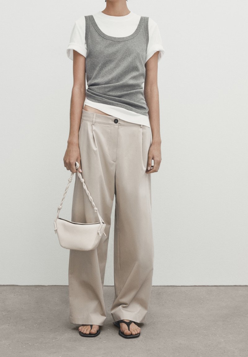 Grijze aansluitende tanktop over een wit T-shirt, beige wijde broek, zwarte flip-flops en een kleine beige handtas met een gevlochten schouderband.