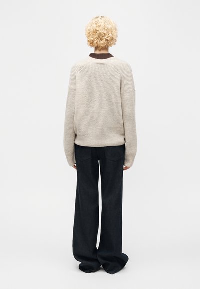 Άτομο με σγουρά ξανθά μαλλιά που φοράει μια φαρδιά μπεζ πλεκτή ζακέτα και φαρδιά μαύρη παντελόνα, στέκεται με την πλάτη γυρισμένη σε απλό φόντο.