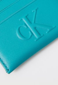 Portofel din piele ecologică de culoare teal, cu logo-ul „CK” embosat, suprafață texturată și mai multe sloturi pentru carduri, cu accente de cusătură ordonate.