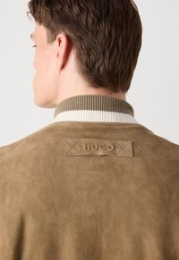 Rjava nubuk jakna z visokim rebrastim ovratnikom, okrašena s kremnimi in olivnimi črtami. Na hrbtni strani je vtisnjen logotip "HUGO". Gladka tekstura.