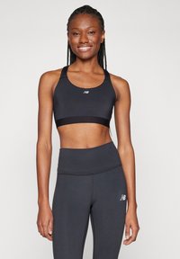 Γυναίκα με μακριές πλεξούδες που χαμογελάει, φορώντας μαύρο αθλητικό σουτιέν και ψηλόμεσα κολάν, στέκεται μπροστά σε απλή ανοιχτόχρωμη παρα背景.