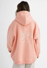 Felpa oversize rosa con cappuccio regolabile. Presenta una linea artistica bianca di un volto e un design di foglia sulla schiena, realizzata in morbido cotone.