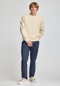 Felpa in cotone beige con testo "Polo Club" in rilievo, abbinata a pantaloni blu e sneaker di colore chiaro. Design semplice e casual.