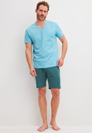 Man wearing light blue short-sleeve V-neck shirt and patterned dark green shorts standing barefoot against white wall.