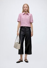 Femme debout portant une chemise rose à manches courtes boutonnée, un pantalon large noir court, des ballerines noires, tenant un sac à main beige.
