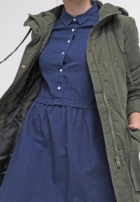 Olivově zelená parka s kapucí a zipy, oblečená přes modrou košilovou šaty s límečkem a skládací sukní.