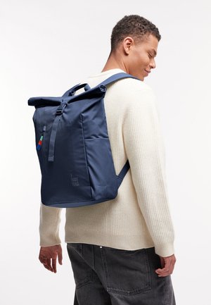 Uomo con maglione crema e jeans scuri con zaino navy blu a roll-top con cerniera frontale e scritta "GOT BAG" su sfondo bianco.