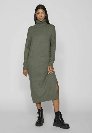 Sweater dress in olive green knit material, featuring a turtleneck, long sleeves, and side slits. Paired with black chunky boots.