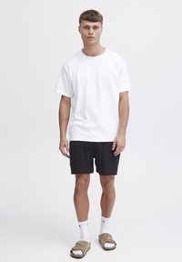 Camiseta blanca de algodón con mangas cortas, combinada con pantalones cortos negros con textura. Unos calcetines blancos y sandalias deslizantes marrones completan el conjunto.