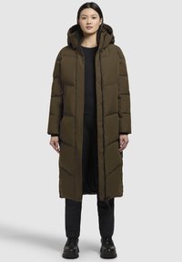 Dlouhá olivově zelená péřová bunda s kapucí, s prošívaným vzorem, předním zipem a postranními knoflíky. Nositá přes černý top a kalhoty.