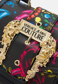 Černá kabelka s vzorem, která má zlatý uzávěr s dekorativními akcenty v podobě lvů a živé květinové vzory v modré, růžové a žluté barvě.