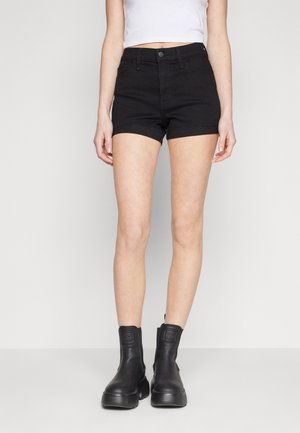 Kvinde iført sorte højtaljede denimshorts, hvid kort top og sorte chunky ankelstøvler stående mod en ensfarvet hvid baggrund.