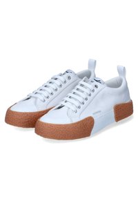 Sneaker bianche in tela con suola in gomma marrone texturizzata, design con lacci e occhielli in metallo. Presenta una linguetta sul retro per facilitare l'indossamento.