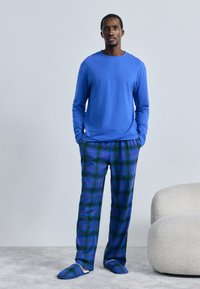 Muž stojí s rukami v vreckách, oblečený v modrej košeli s dlhým rukávom, modro-zelených károvaných nohaviciach a zodpovedajúcich papučiach vedľa bieleho kresla.