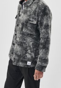 Mężczyzna ubrany w szaro-czarną teksturowaną kurtkę zapinaną na guziki z kieszeniami na klatce piersiowej i białą metką przy dolnym obszyciu, w połączeniu z czarnymi spodniami.