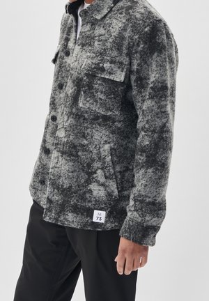 Homme portant une veste boutonnée texturée grise et noire avec des poches poitrine à rabat et une étiquette blanche près de l'ourlet, associée à un pantalon noir.