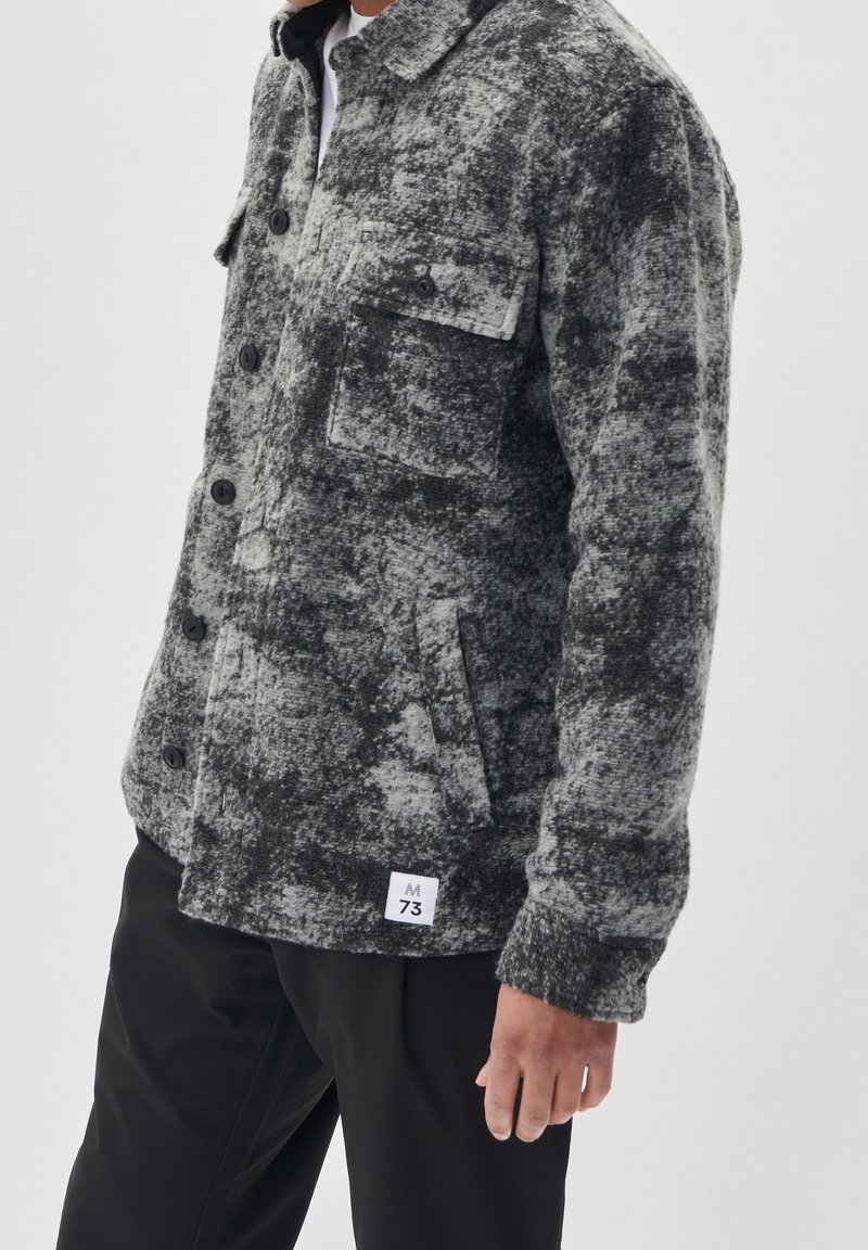 Mężczyzna ubrany w szaro-czarną teksturowaną kurtkę zapinaną na guziki z kieszeniami na klatce piersiowej i białą metką przy dolnym obszyciu, w połączeniu z czarnymi spodniami.