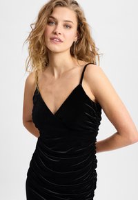 Schwarzes Samtkleid mit V-Ausschnitt und dünnen Trägern. Es hat eine geraffte Textur an den Seiten für eine taillierte Silhouette. Glatte Stoffoberfläche.