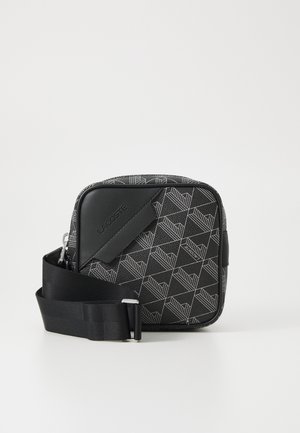 Sac bandoulière noir Lacoste avec sangle ajustable, motif géométrique et logo de la marque embossé sur le panneau avant.