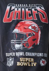 Črna majica s kratkimi rokavi, ki ima na sebi rdečo nogometno čelado z logotipom, drzne besede "KANSAS CITY CHIEFS" in podrobnost "SUPER BOWL CHAMPIONS 1969".
