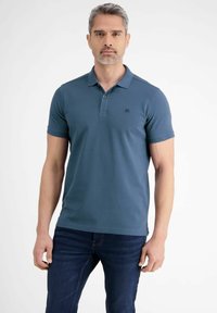 Koszulka polo w ciemnym teal z prążkowanym kołnierzykiem i krótkimi rękawami. Posiada subtelne logo na piersi. W zestawie z ciemnoniebieskimi dżinsami.