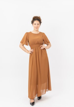 Jeune femme aux cheveux bouclés, portant une longue robe marron et des chaussures noires, debout les mains sur les hanches sur fond blanc.
