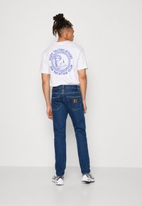 Hvit t-skjorte med blå grafikk på baksiden, kombinert med mørkeblå jeans. Antrekket inkluderer hvite og svarte sneakers med teksturert design.