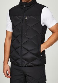 Černá prošívaná vesta s vysokým límcem, předním zapínáním na zip a bočními kapsami. Má texturovanou látku a elegantní design. Nositelná přes bílou košili.