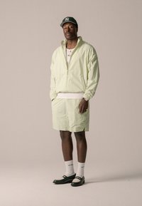 Veste zippée texturée vert clair et shorts assortis. Chemise blanche en dessous. Casquette verte, chaussettes blanches et mocassins bicolores.