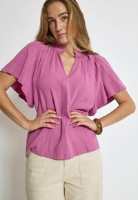 Top rosa a maniche corte con scollatura a V e dettagli arricciati sul collo. Indossato con pantaloni chiari a vita alta. Tessuto liscio.