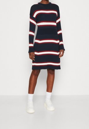 Person trägt ein dunkelblaues Kleid mit weißen und roten horizontalen Streifen, kombiniert mit weißen Socken und weißen Sneakers, steht vor einem weißen Hintergrund.