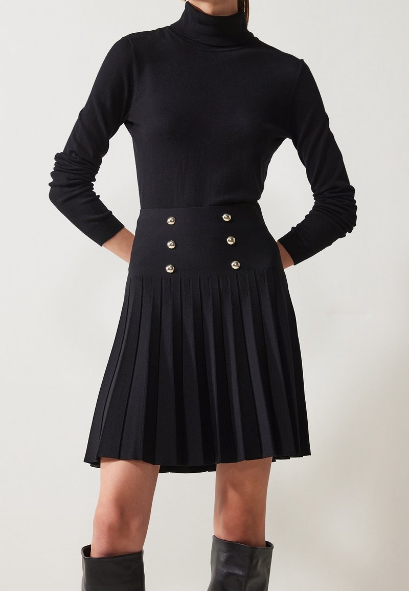 Femme portant un haut noir à col roulé à manches longues et une jupe plissée noire avec six boutons dorés, associée à des bottes noires montant jusqu'aux genoux.