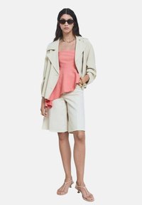 Blazer oversize beige, top peplum corallo, pantaloni corti crema e sandali nudi. Gli accessori includono un braccialetto dorato e occhiali da sole neri.