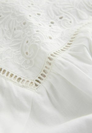 Weißer Stoff mit aufwändigen Stickmustern, die entlang des Randes ausgeschnittene Details aufweisen, sowie einer weichen, strukturierten Oberfläche. Leicht und zart.